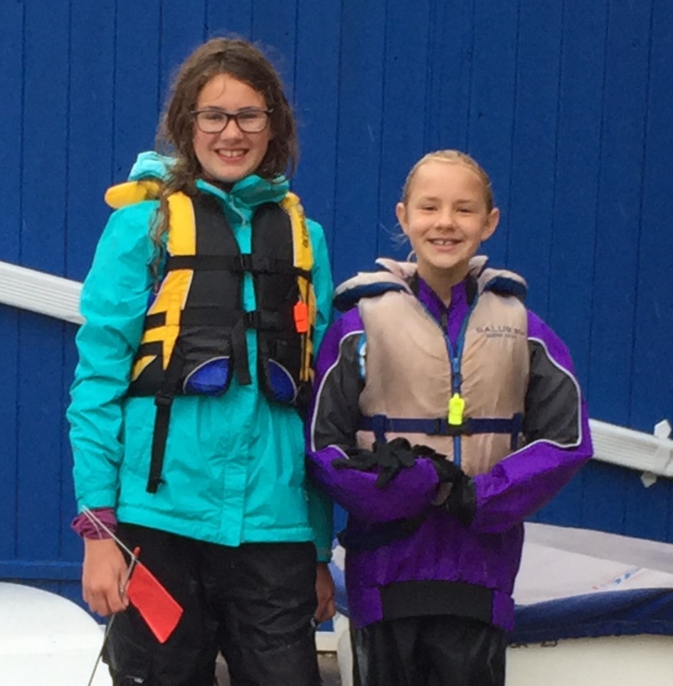 Annie as a young sailor in a life jacket and rain gear, standing beside Optimist dinghies in the rain.